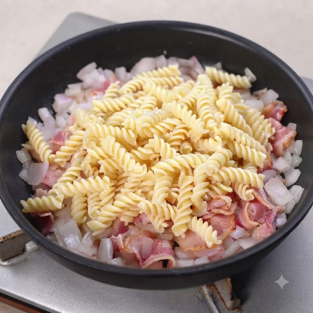 Pasta en espiral cocida descansando sobre tocino y cebollas cocinadas en una sartén negra.
