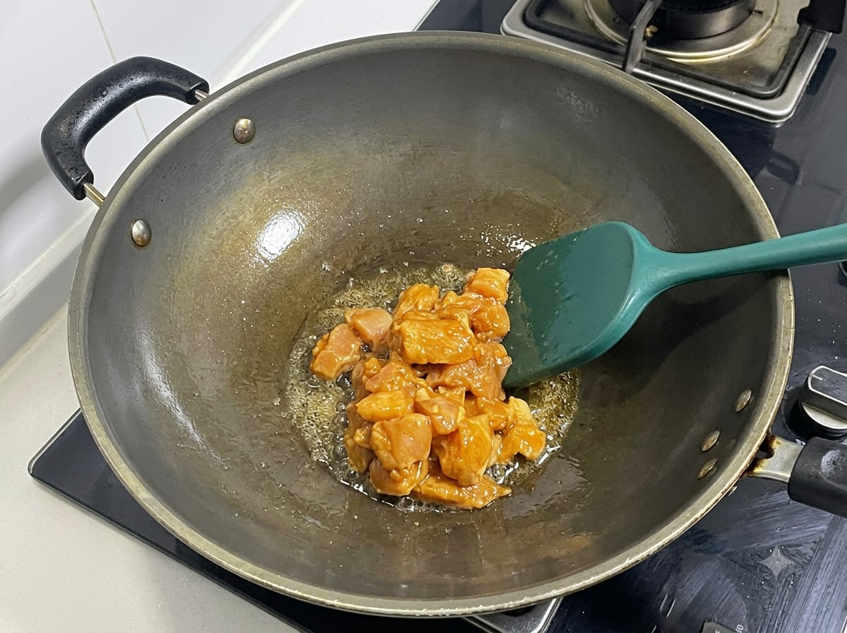 Pechuga de pollo marinada en cubos siendo salteada en un wok caliente con una espátula verde.