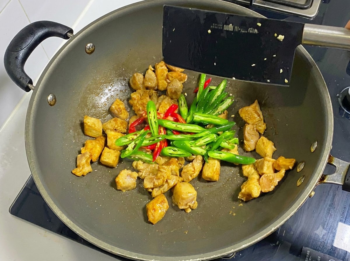 Chiles verdes y rojos en rodajas añadidos a los trozos de pollo en un wok, mezclados con una espátula negra.