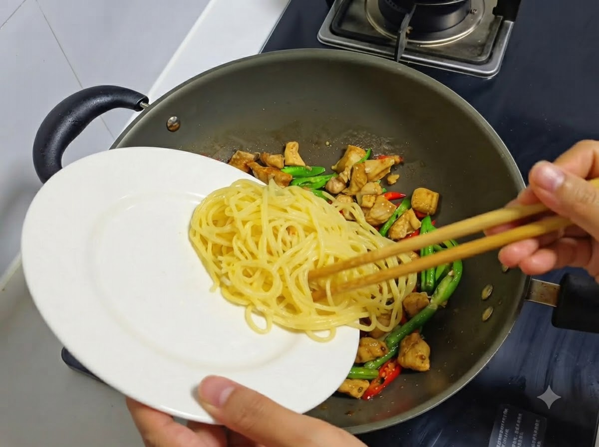 Espaguetis cocidos siendo transferidos de un plato blanco a un wok que contiene pollo y chiles.