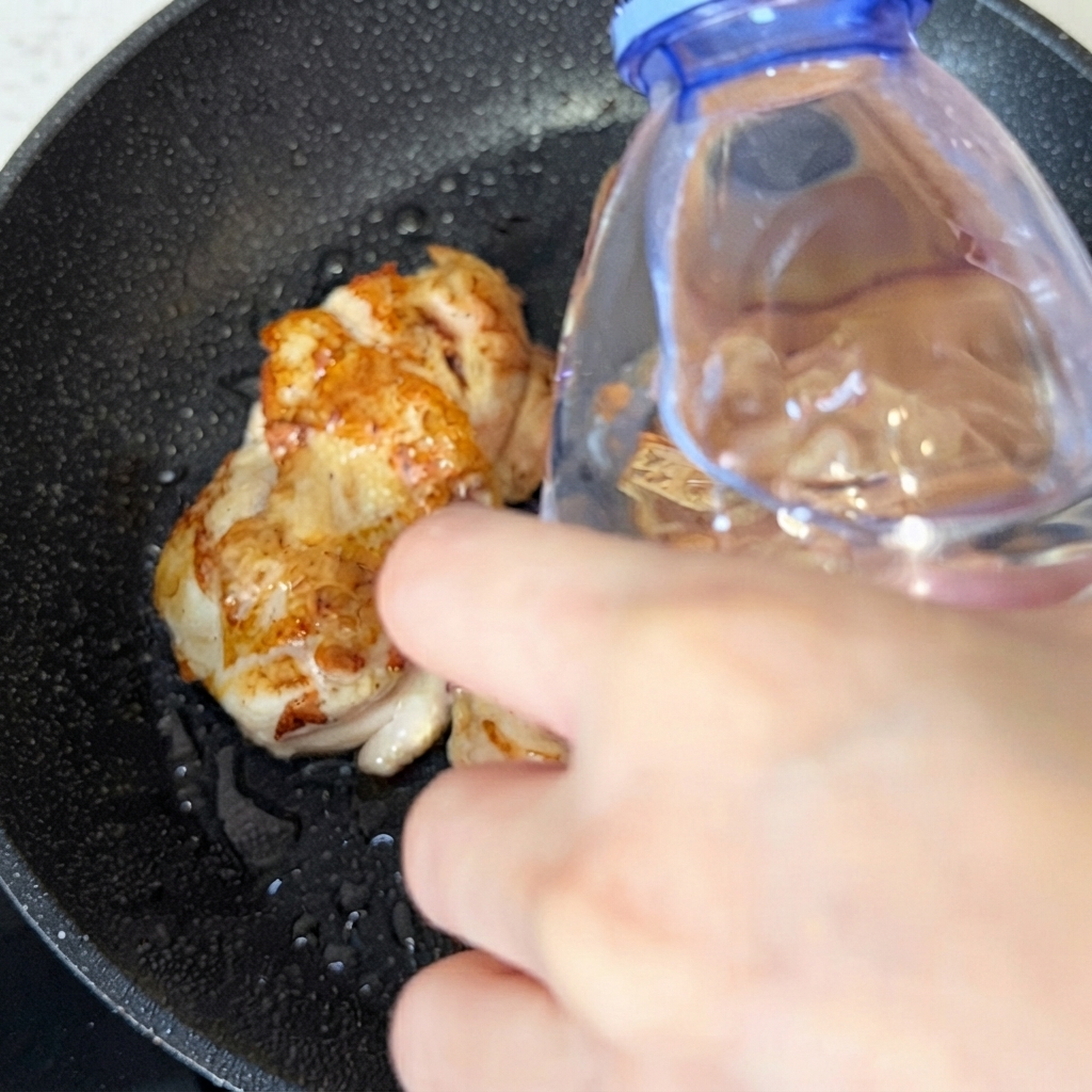 Una botella vertiendo un poco de agua en la sartén sobre los trozos de pollo cocinado.