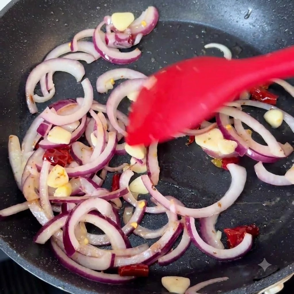 Cebollas rojas, ajo y chiles secos siendo salteados en una sartén caliente con una espátula de silicona roja.