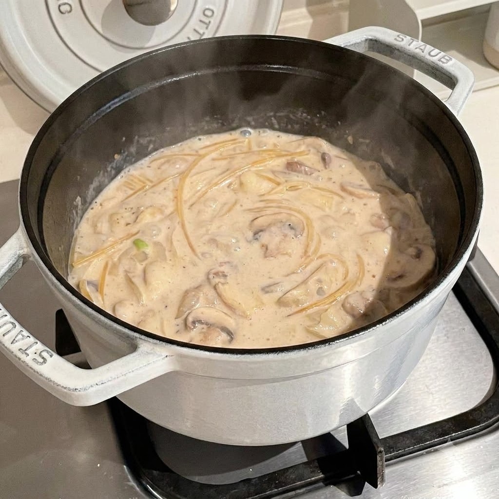 Fideos de espagueti cociéndose dentro de un caldo cremoso hirviendo a fuego lento dentro de una olla blanca.