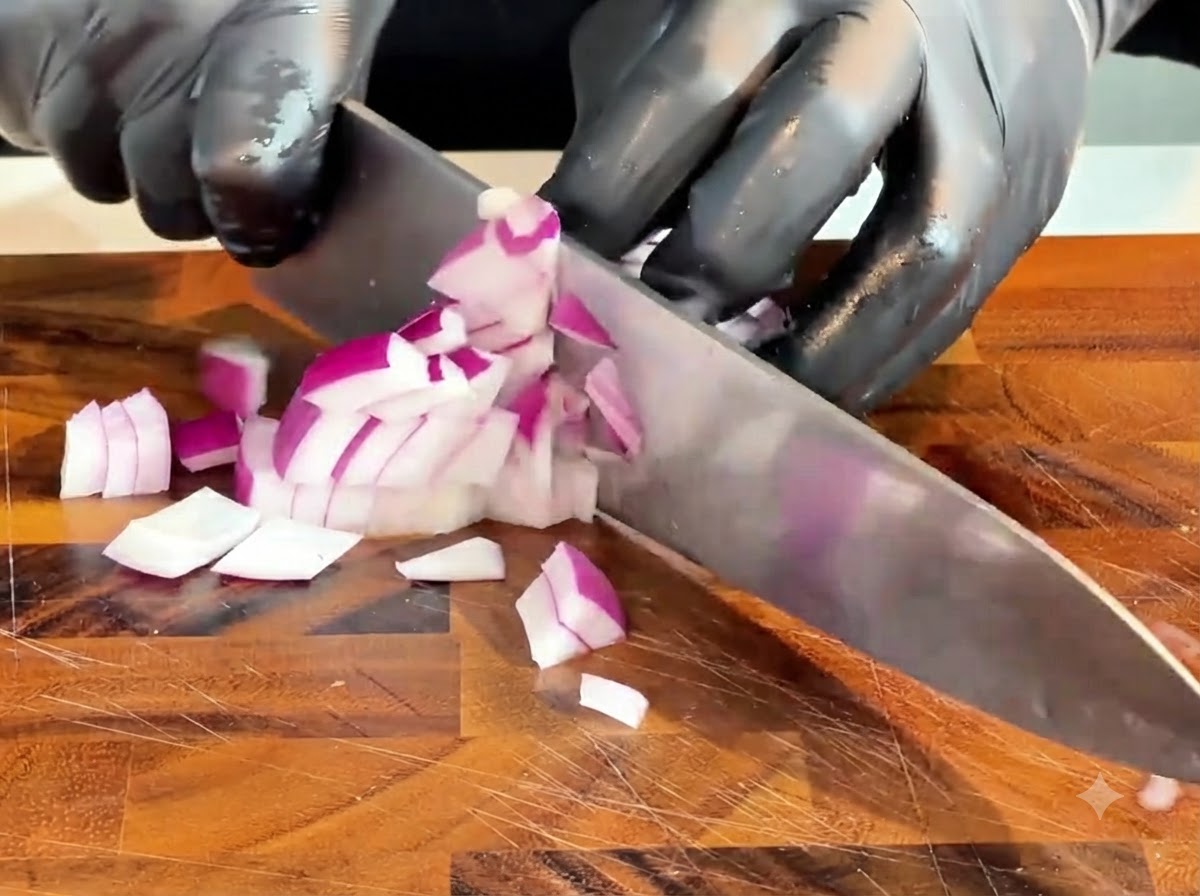 Un cuchillo picando una cebolla morada cruda en trozos pequeños sobre una tabla de cortar de madera.