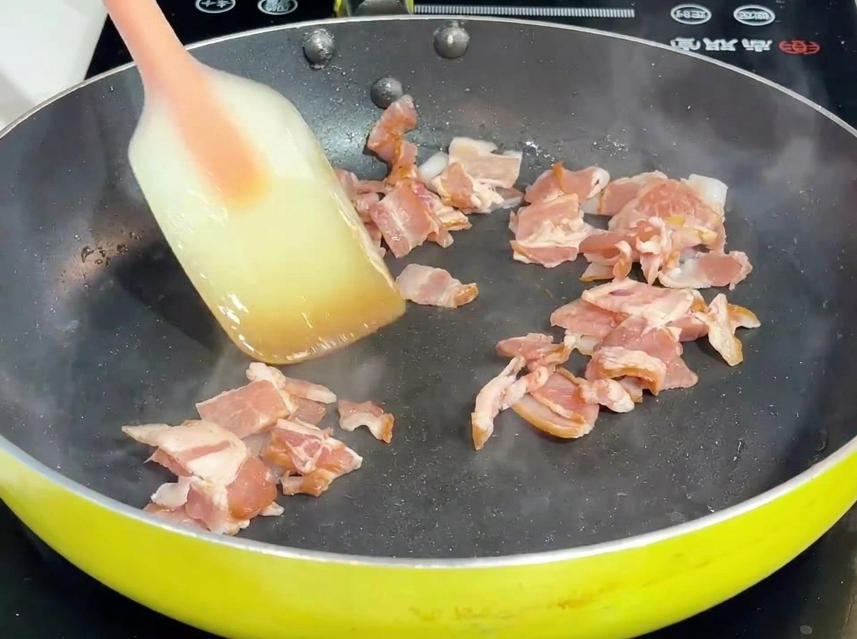 Tocino y cebolla siendo sofritos con una espátula en un sartén.