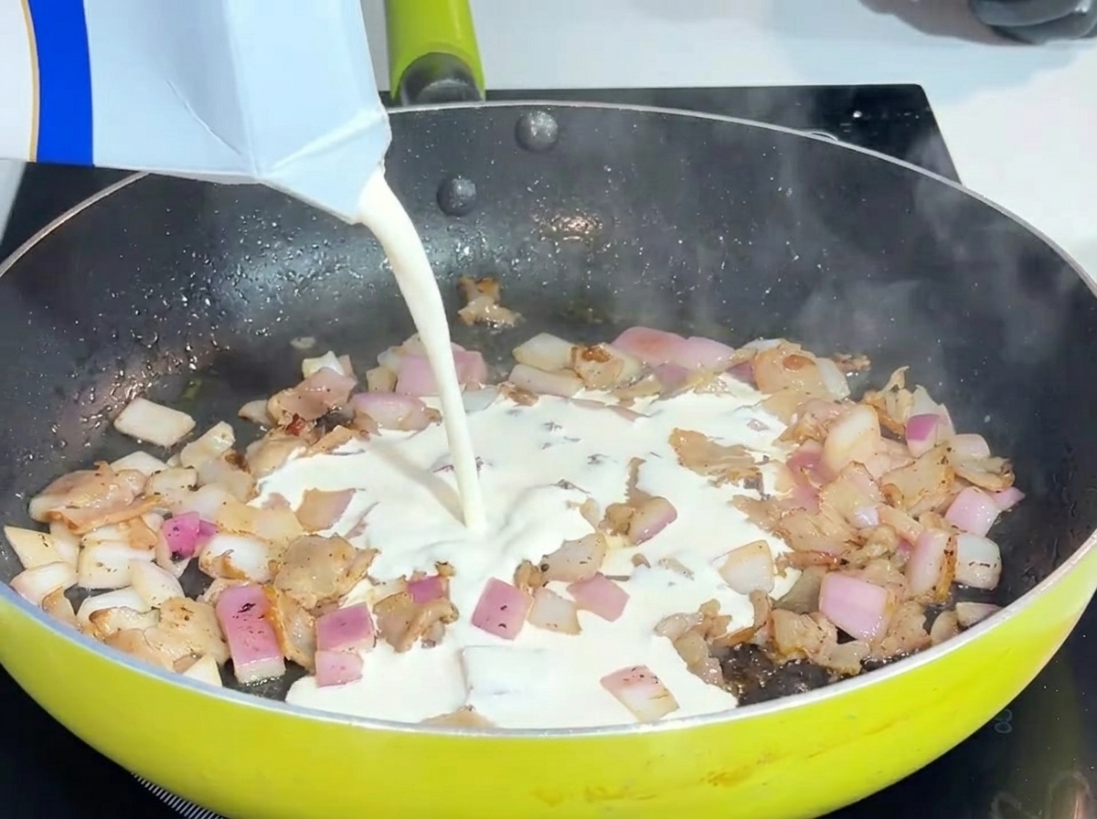 Crema de leche líquida siendo vertida en un sartén con tocino y cebollas.