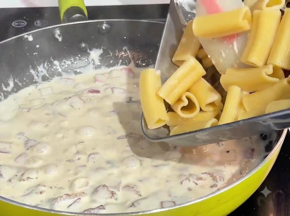 Pasta en forma de corazón pre cocida siendo vertida desde una bandeja en una salsa burbujeante de crema y tocino en un sartén.