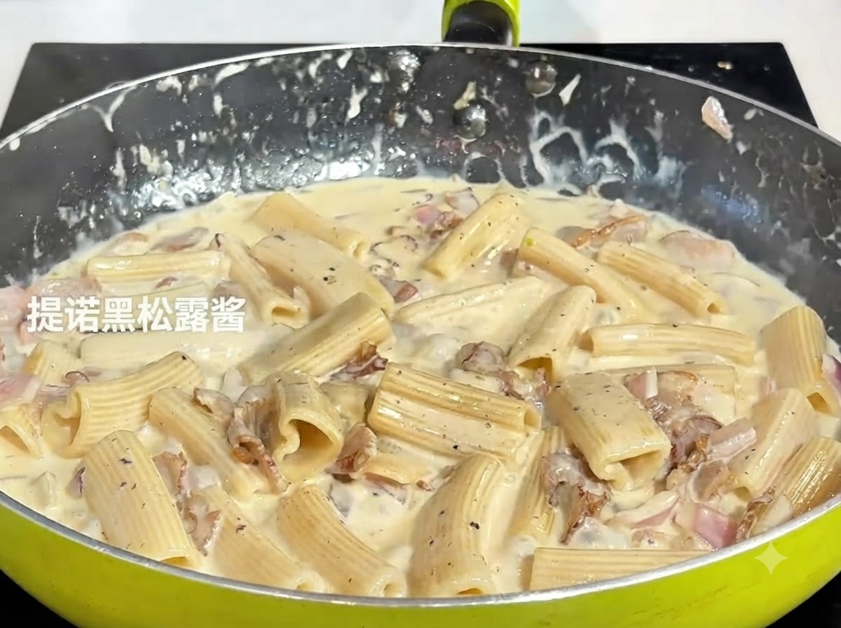 Una cuchara de pasta de trufa negra oscura siendo añadida a la mezcla de pasta a fuego lento en un sartén.