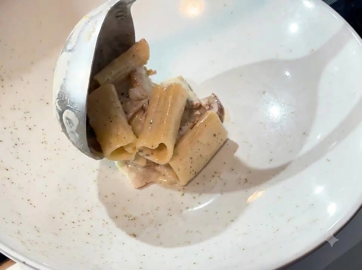 Un cucharón transfiriendo pasta cremosa de un sartén a un plato hondo blanco.