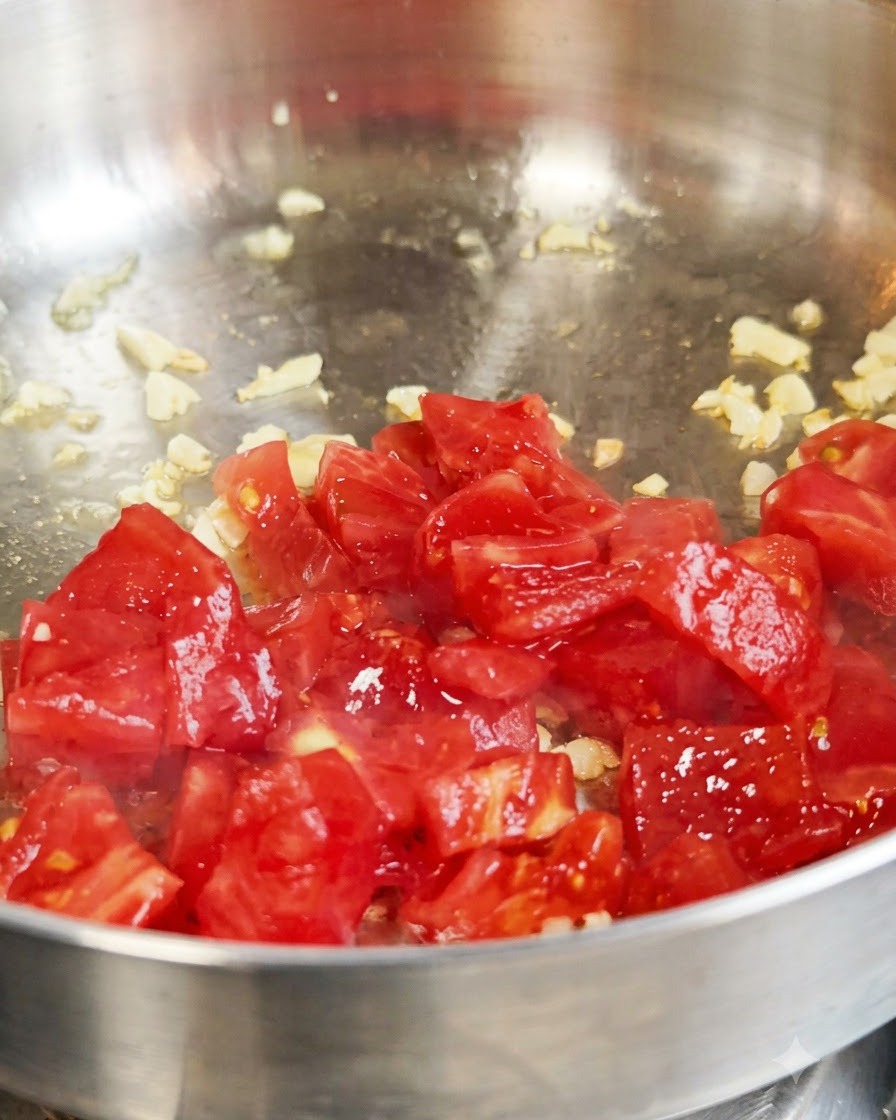 Tomates rojos frescos en daditos salteándose con ajo dentro de una sartén metálica.