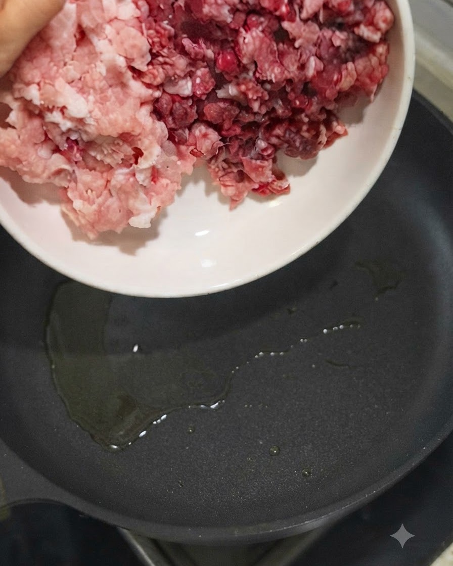 Carne de cerdo y carne de res picada siendo vertidas desde un tazón blanco en una sartén caliente con aceite.
