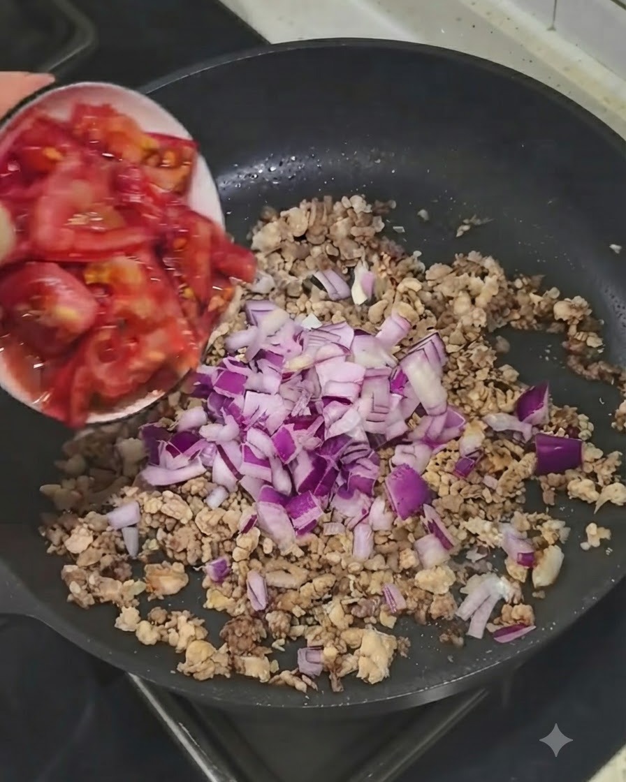 Tomates frescos picados siendo añadidos desde un tazón pequeño a la sartén con carne y cebollas.