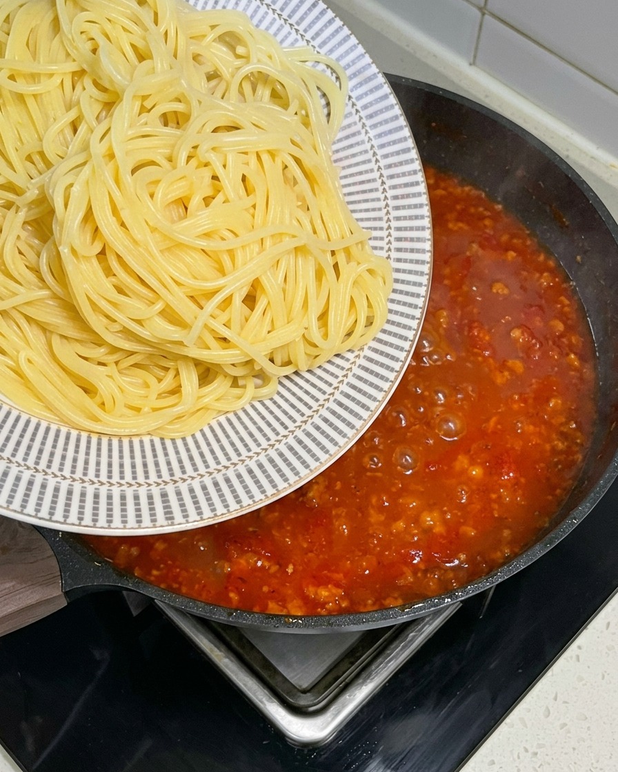 Fideos espagueti recién hervidos siendo transferidos desde un plato estampado a una sartén grande llena de salsa de carne roja burbujeante.