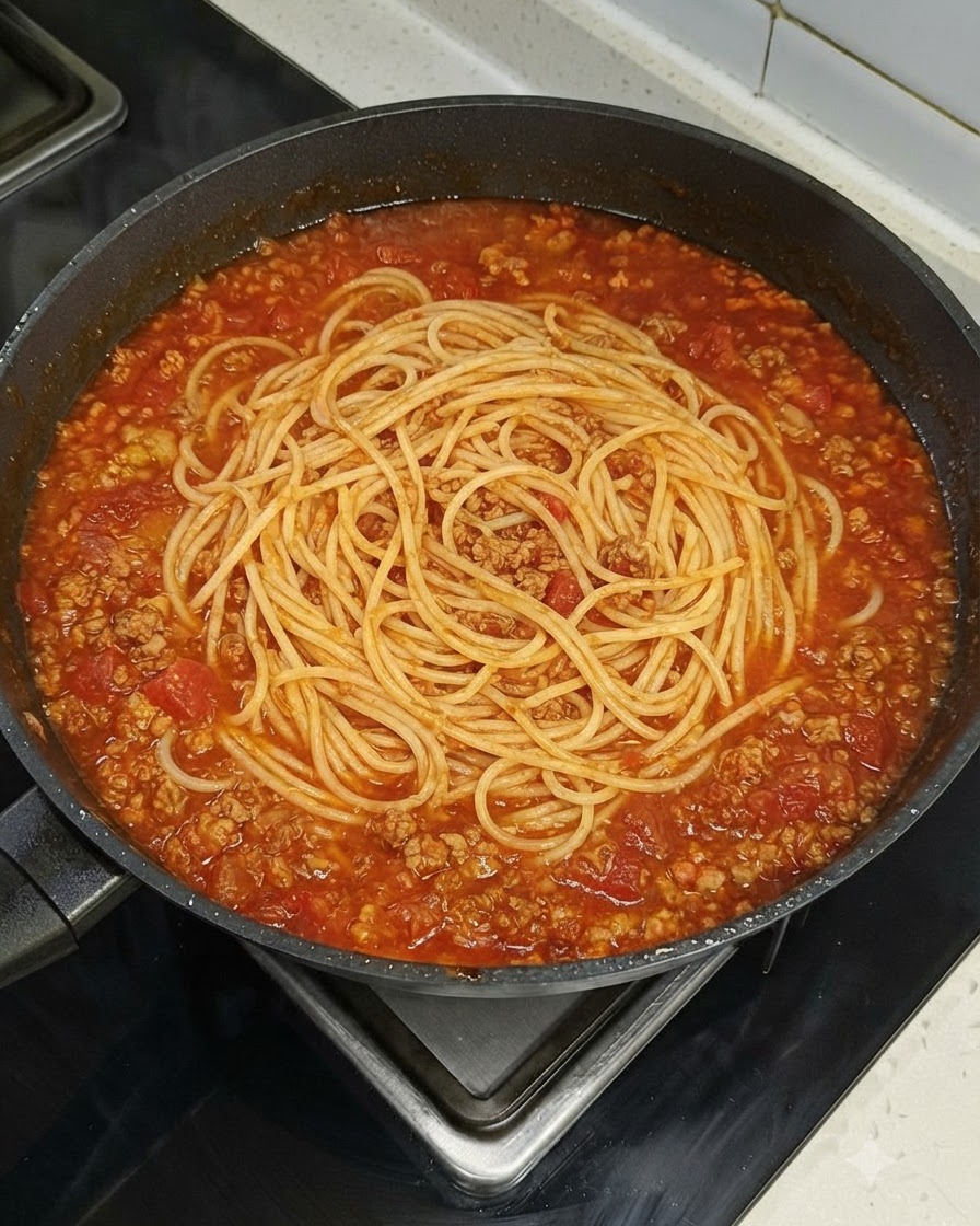 Un nido de fideos espagueti reposando uniformemente dentro de una sartén grande llena de salsa de carne y tomate en una estufa.