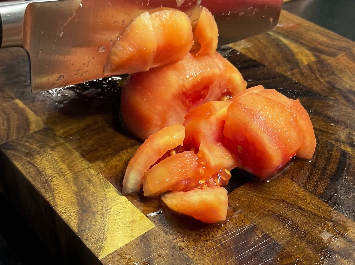 Un cuchillo de cocina picando tomates cocidos y pelados en trozos sobre una tabla de cortar de madera.