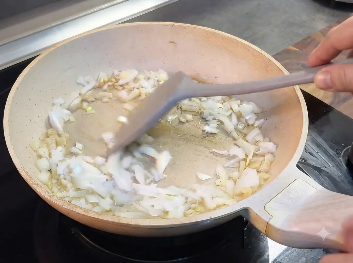 Cebollas picadas siendo revueltas con una espátula gris dentro de una sartén de color claro.