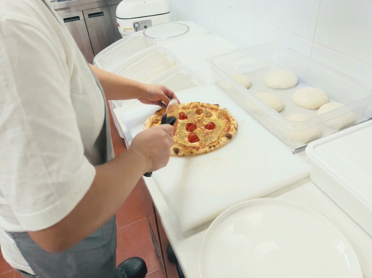 Una persona cortando una pizza recién horneada en porciones con un cortador giratorio sobre una tabla blanca.