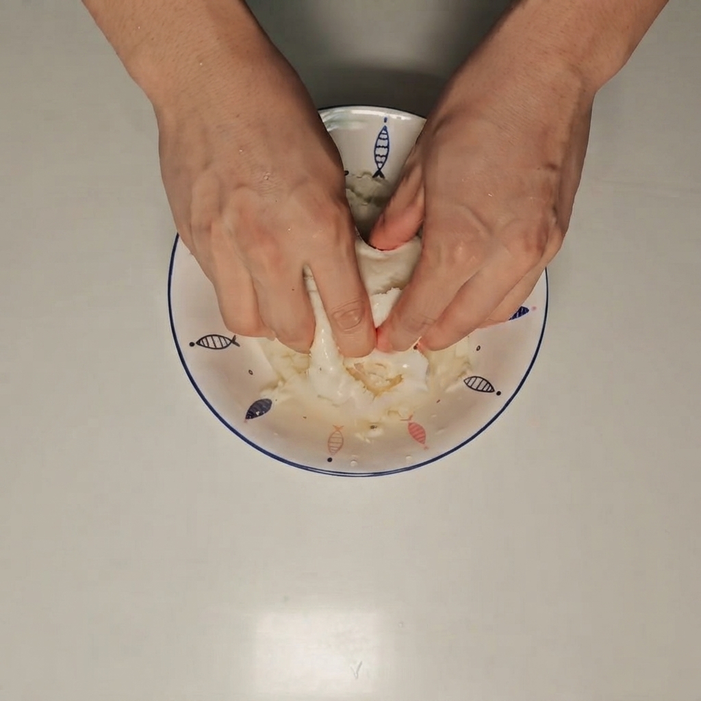 Manos triturando un pan al vapor húmedo dentro de un bol blanco.