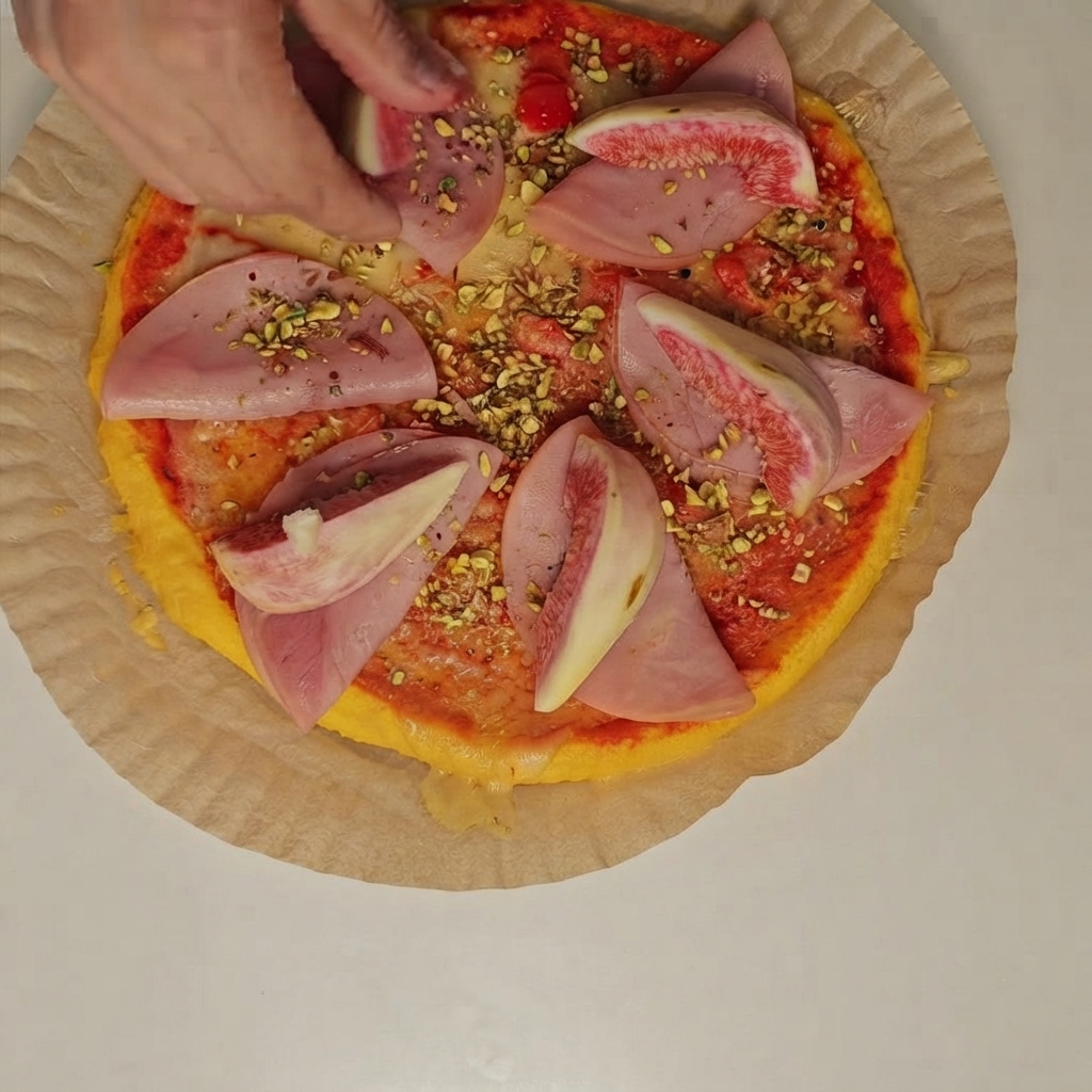Una persona colocando gajos de higos frescos y pistachos picados sobre las rodajas de jamón en la pizza.