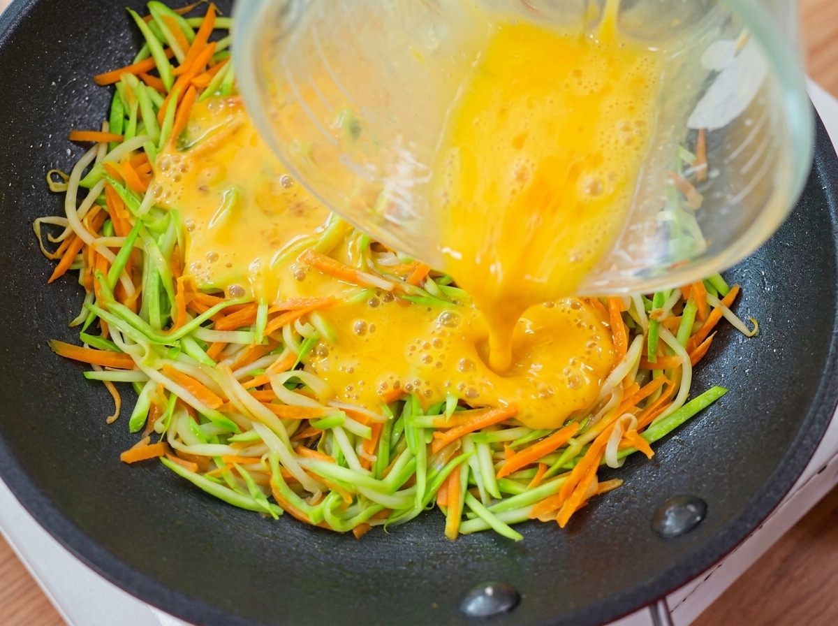 Líquido de huevo amarillo vertiéndose desde una taza medidora sobre la capa de verduras en la sartén.