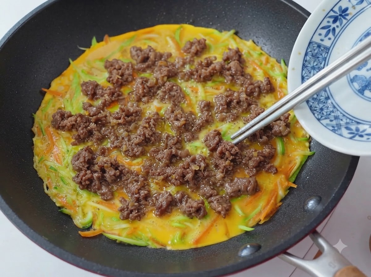 Primer plano de palillos colocando y esparciendo migas de carne cocida sobre una sartén con una base de panqueque de verduras y huevo.