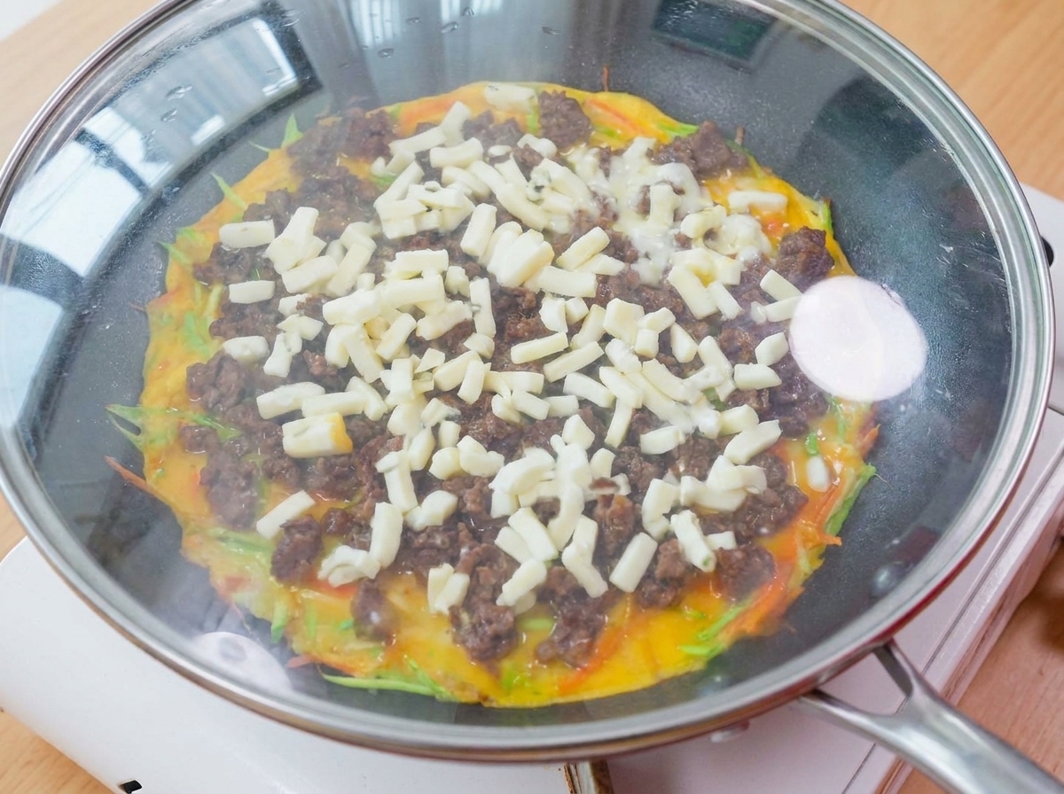 Una sartén cubierta con una tapa de vidrio sobre una estufa, con vapor y queso derretido visible en el interior.