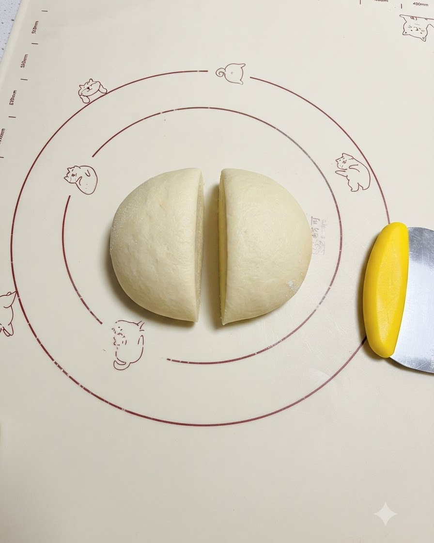 Masa fermentada cortada uniformemente a la mitad sobre una estera de silicona con diseño junto a una espátula de masa amarilla.
