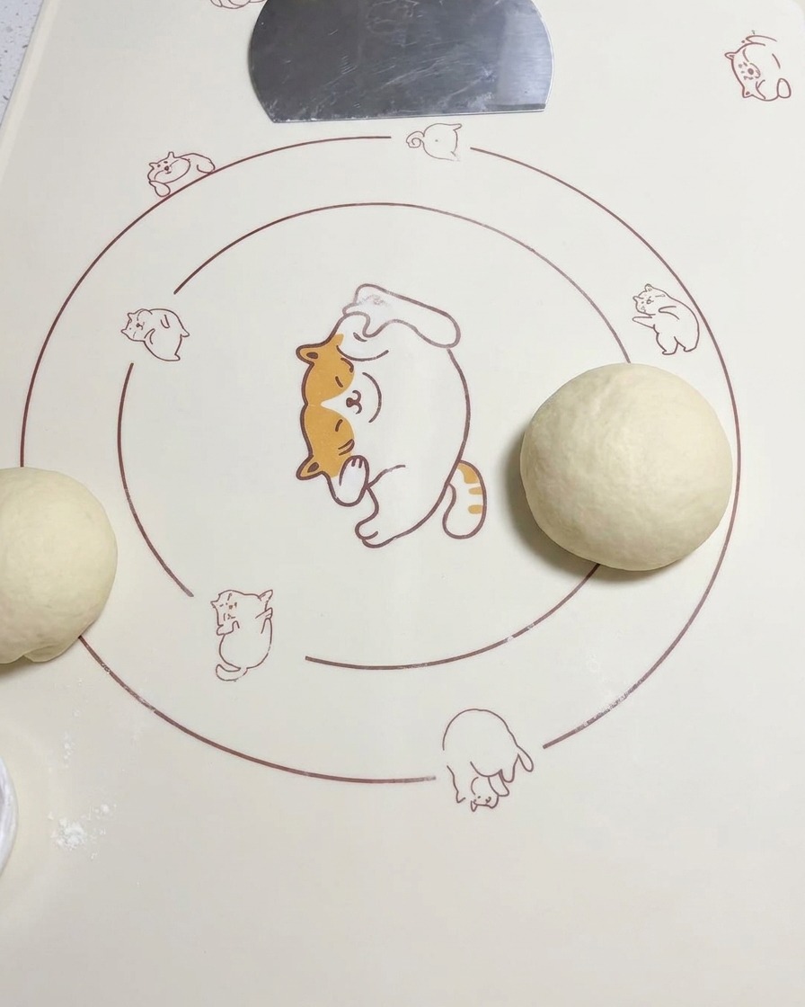 Una bola de masa redondeada colocada sobre una estera de silicona junto a otra pieza de masa estirada.