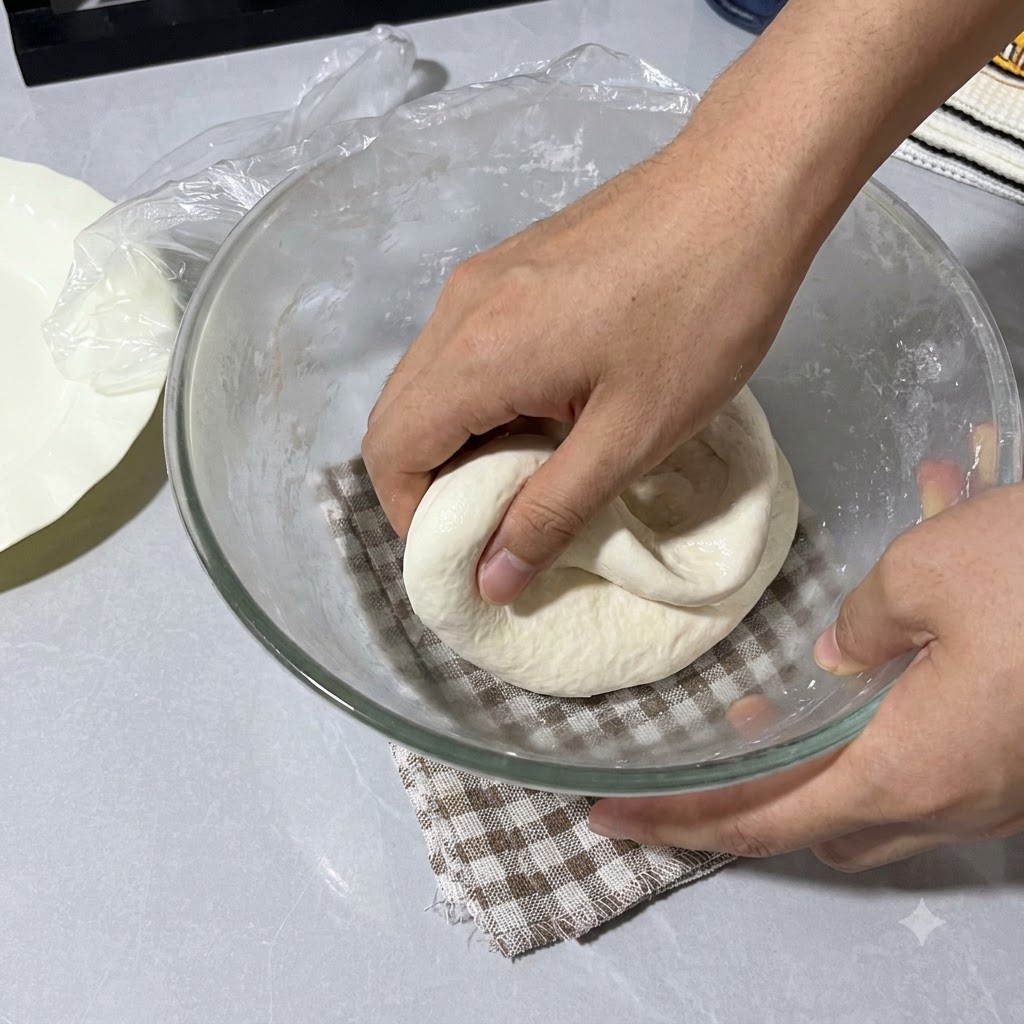 Manos rociadas con agua doblando la masa dentro del tazón de vidrio cubierto con papel film.