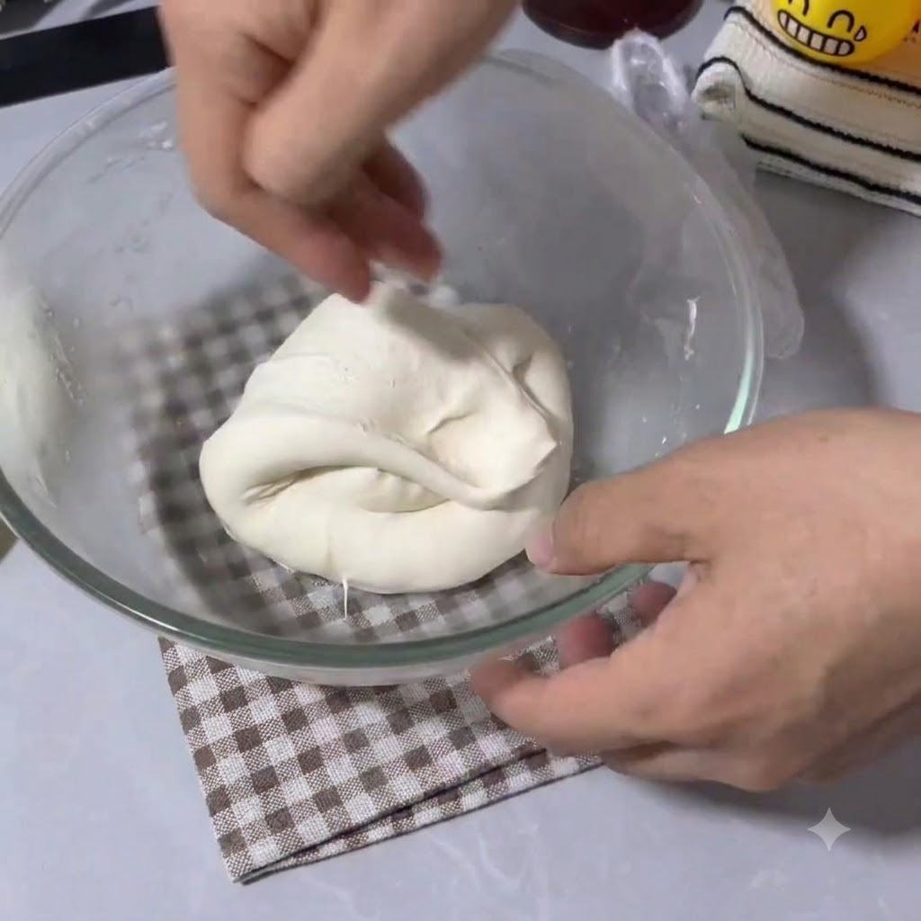 Manos doblando y estirando masa elástica de pizza dentro de un tazón de mezcla de vidrio transparente.