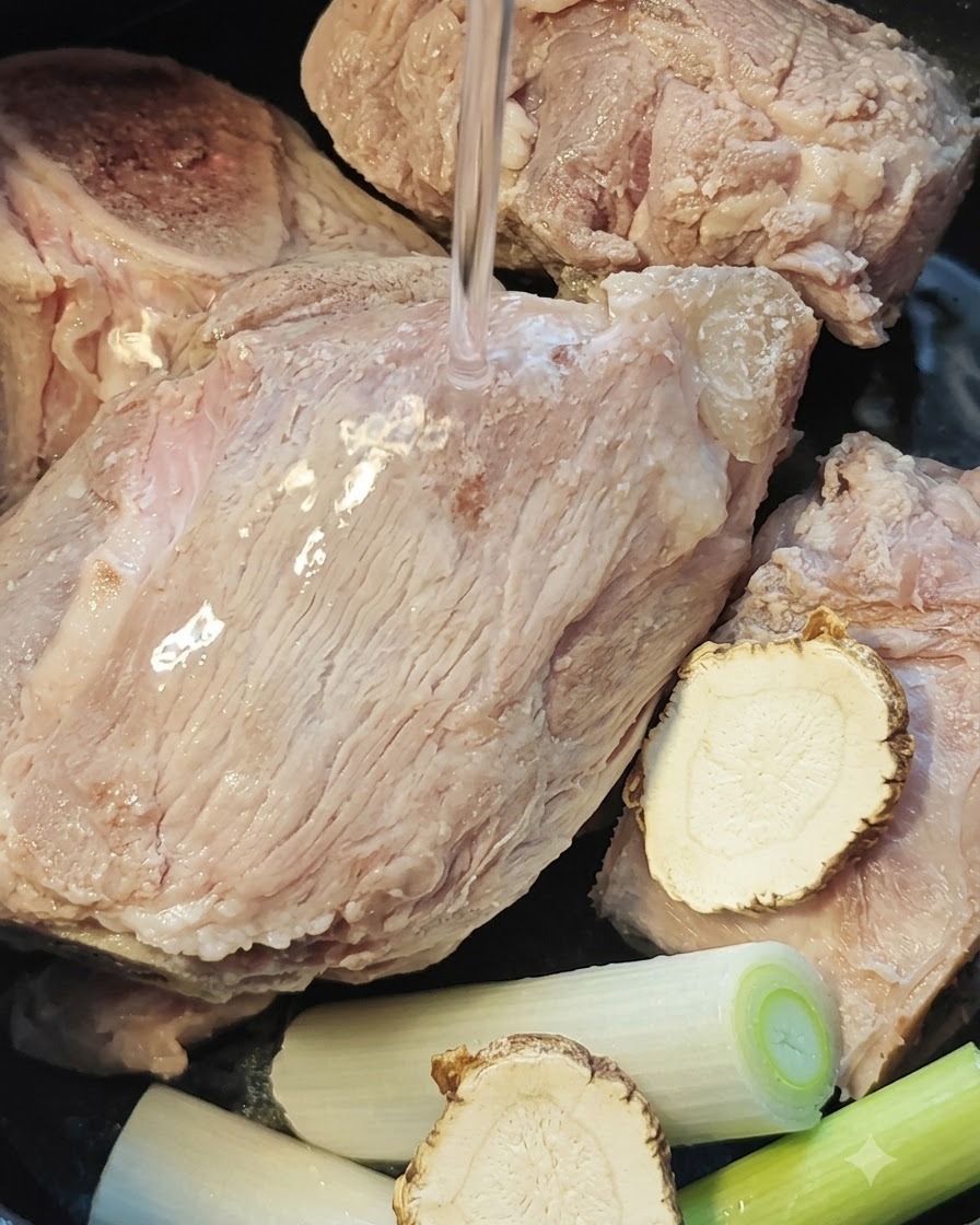 Agua siendo vertida sobre los huesos de cerdo, la raíz de angélica y los cebollinos dentro de la olla de una arrocera.