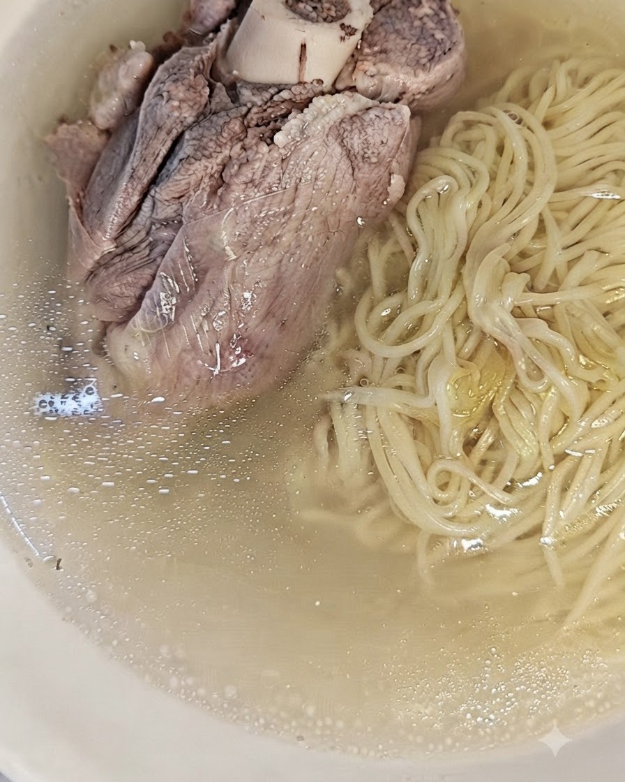 Fideos cocidos reposando en un tazón junto al caldo de cerdo claro y colado.