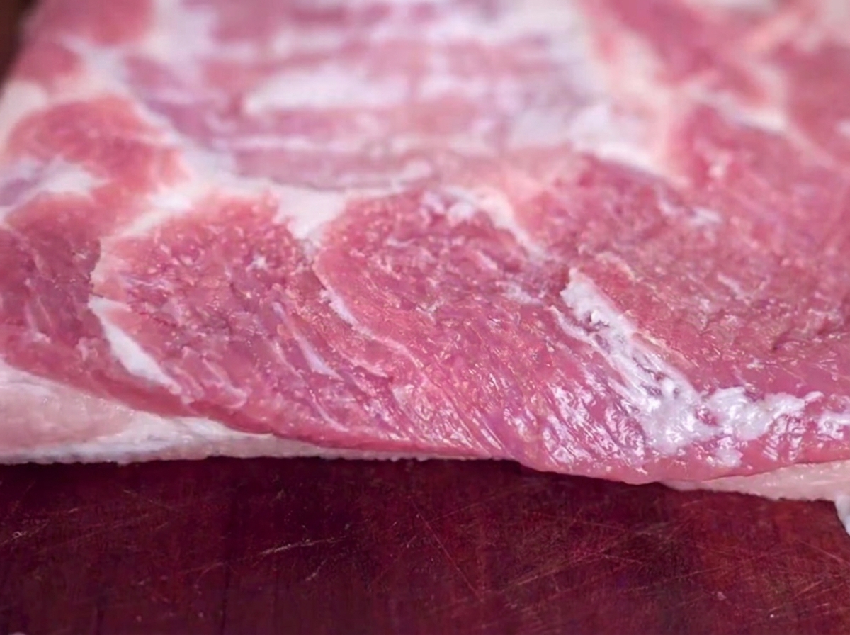 Primer plano de un trozo de panceta de cerdo cruda sobre una tabla de cortar de madera con granos de sal visibles.