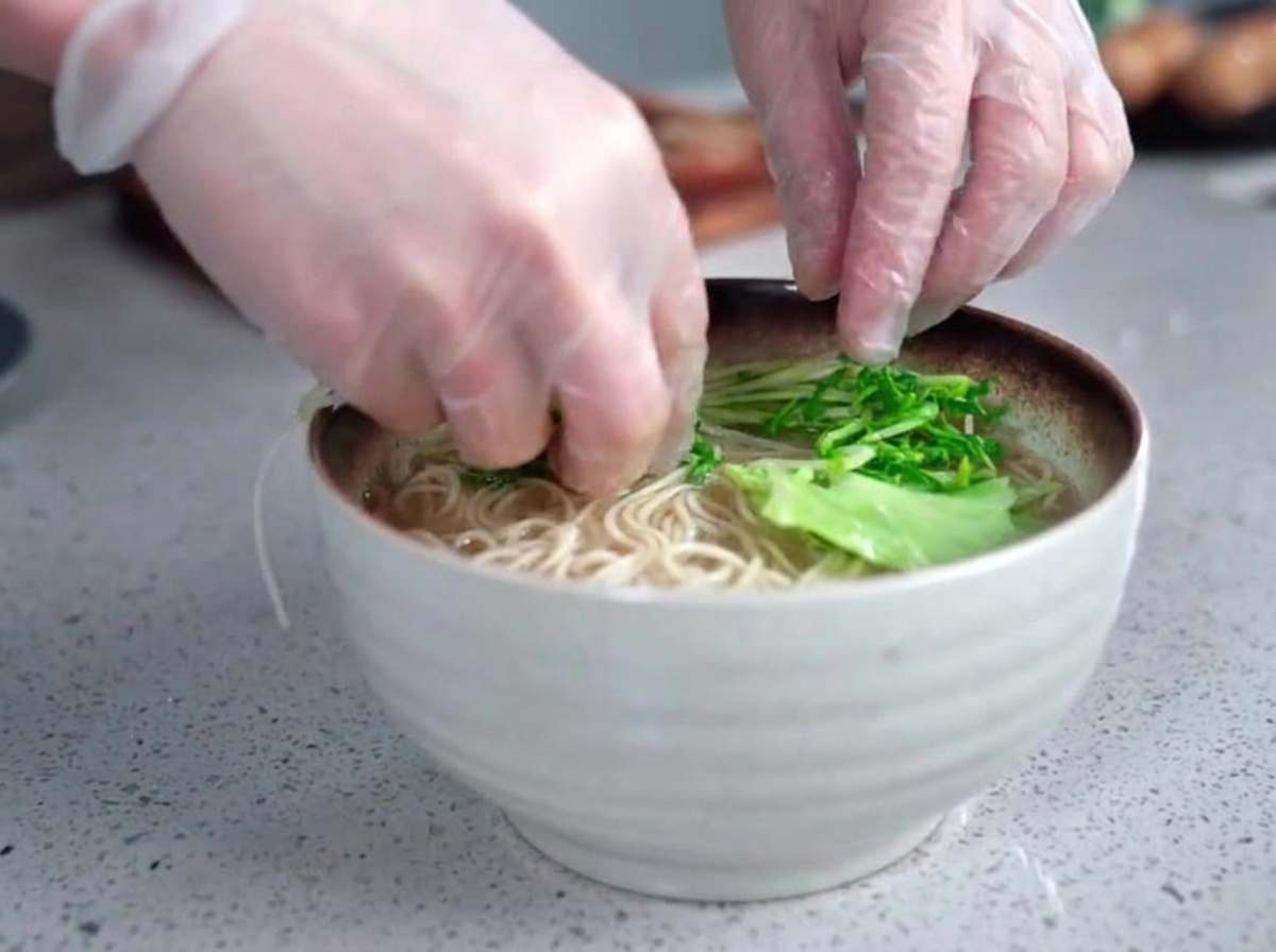 Manos con guantes colocando cuidadosamente verduras de hoja verde fresca en un tazón humeante de fideos de ramen y caldo opaco.