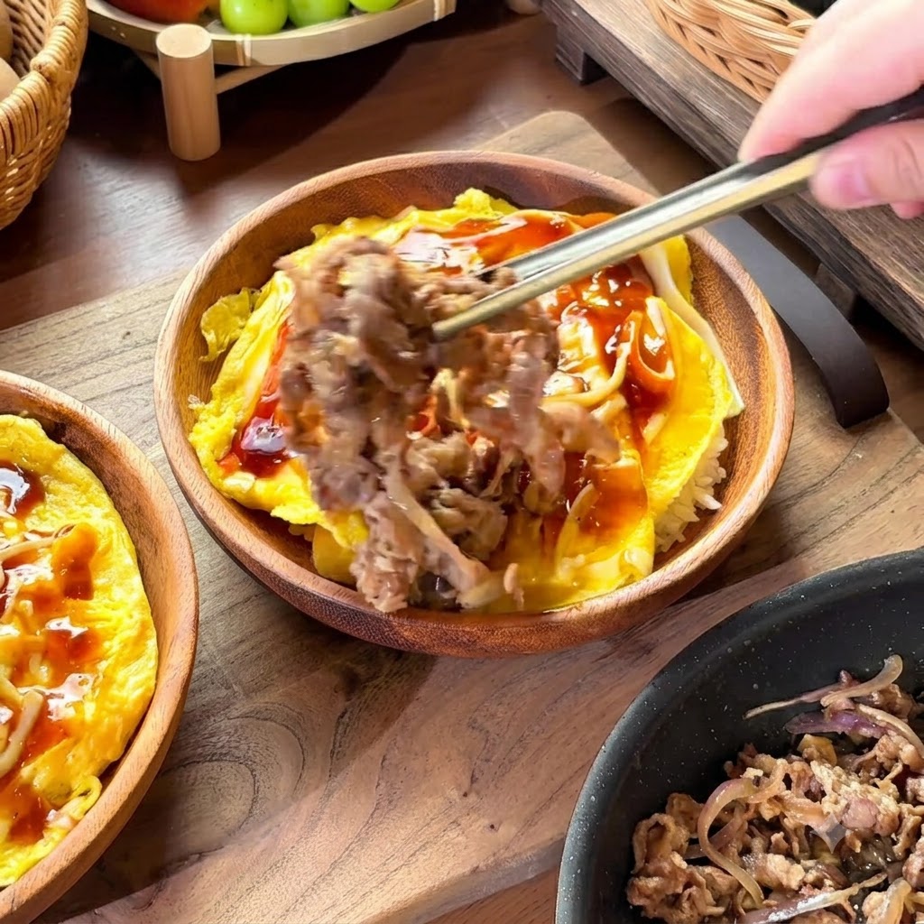 Palillos levantando una porción de ternera cocida y cebollas para colocarla sobre un tazón de arroz y huevos cubiertos de salsa.