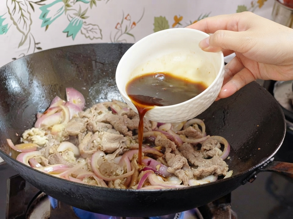 Salsa siendo vertida desde un pequeño bol blanco en un wok con cebollas y trozos de ternera.