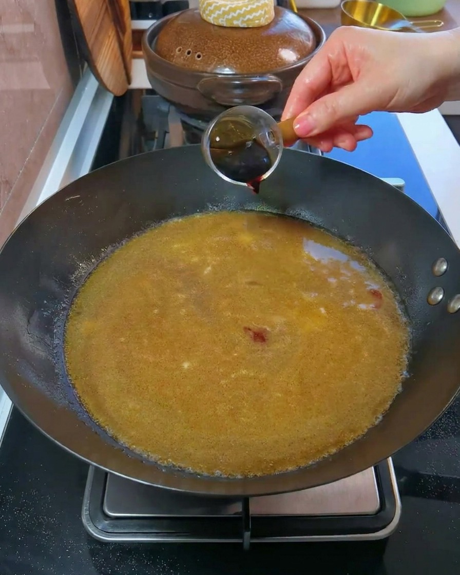 Una mano vertiendo salsa de soja oscura desde un vaso medidor pequeño en un wok lleno de caldo.
