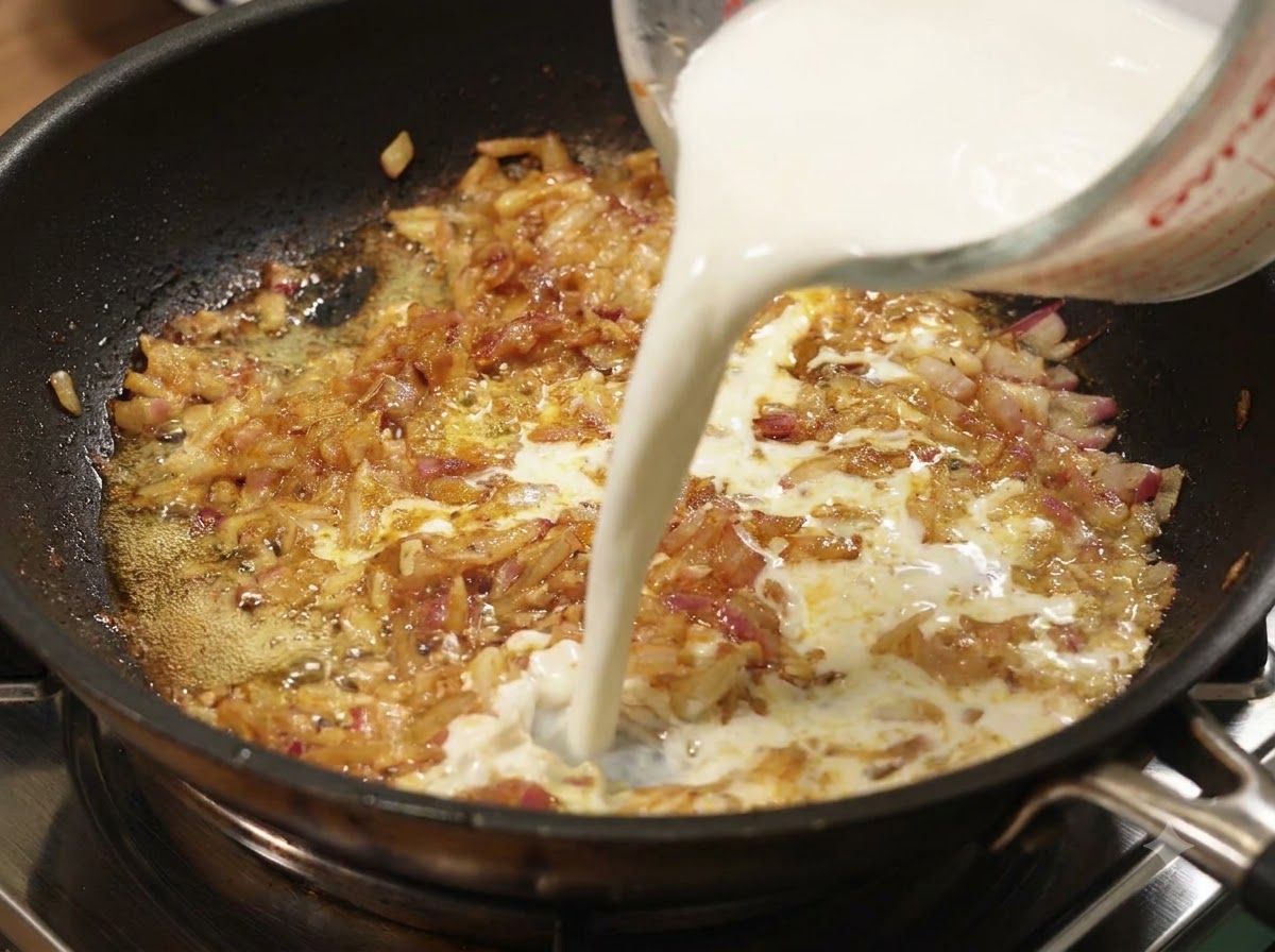 Leche siendo vertida desde una taza medidora en una sartén con cebollas y curry a fuego lento.
