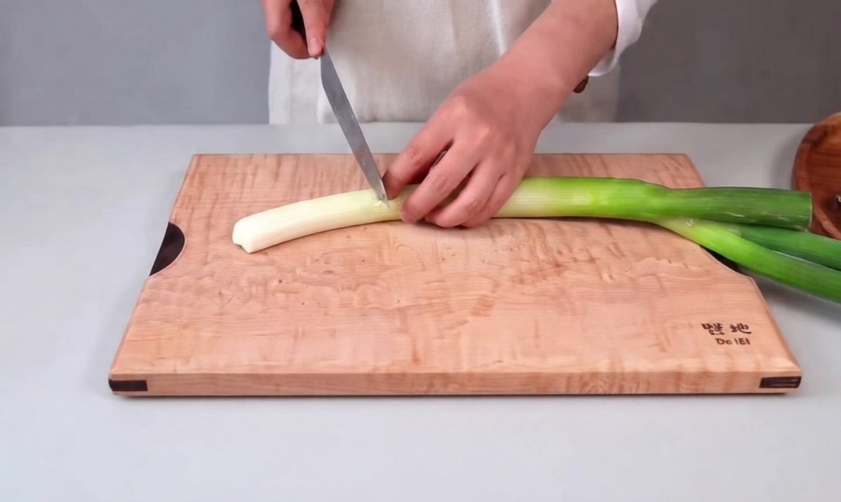 Manos sosteniendo un cuchillo de cocina y cortando una cebolleta grande en una tabla de madera.