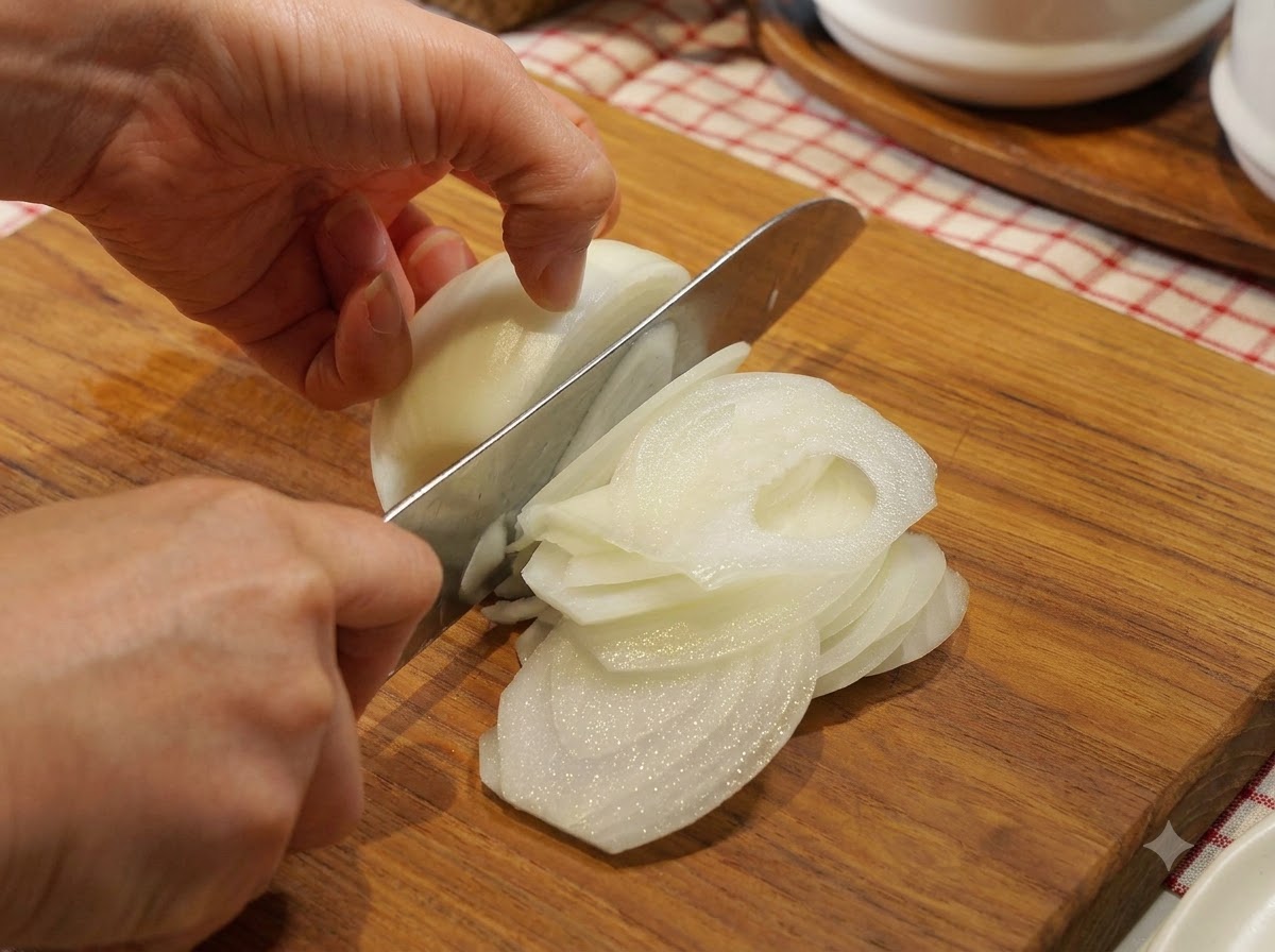 Una cebolla siendo cortada cuidadosamente en medias lunas finas sobre una tabla de cortar de madera con un cuchillo de chef.