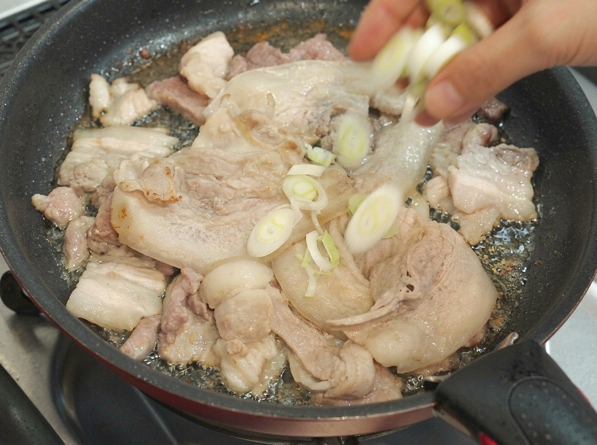 Cebollas verdes picadas cayendo en una sartén que contiene rodajas de panceta de cerdo en cocción.