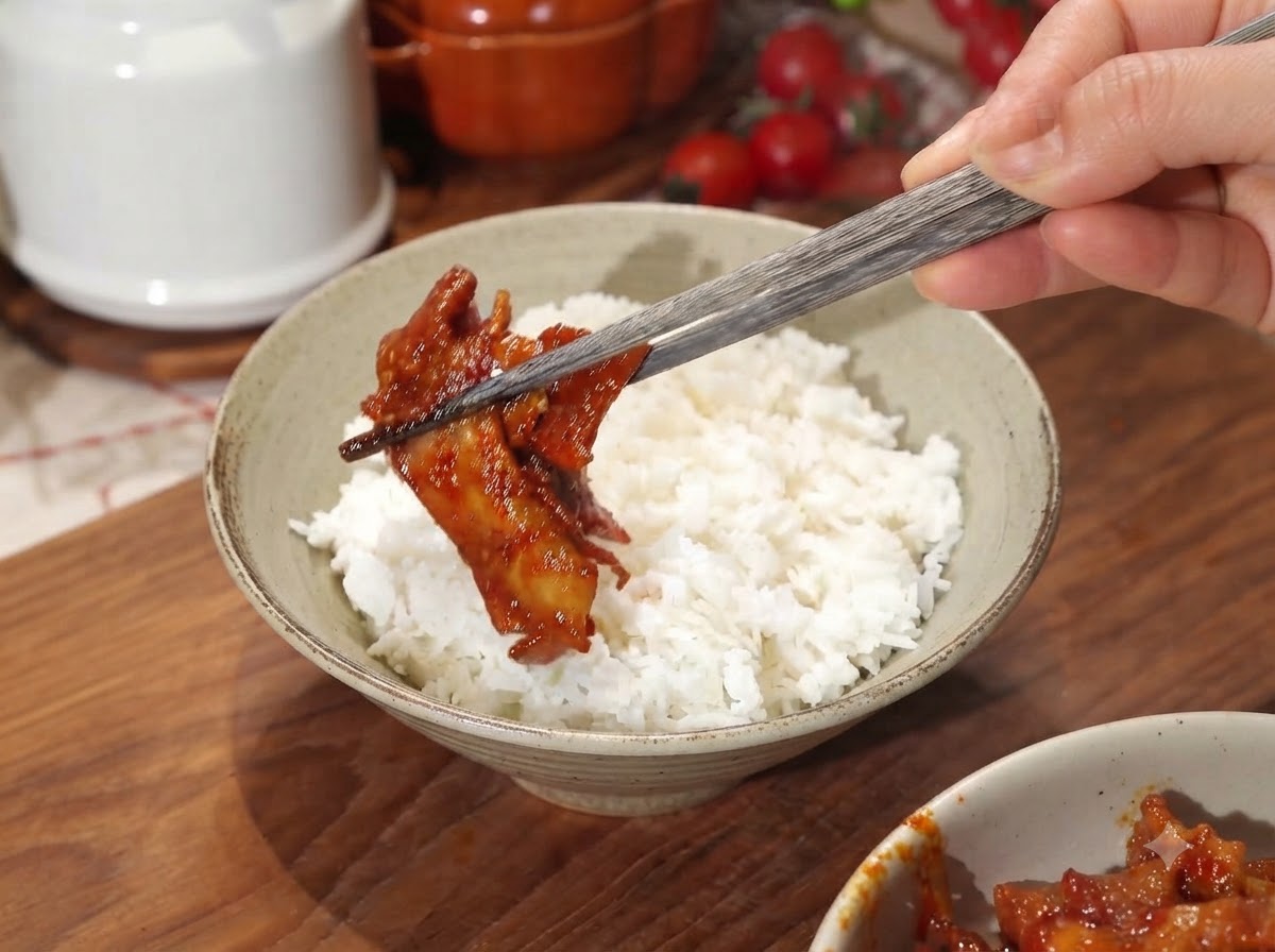 Una mano sosteniendo palillos, colocando un trozo de cerdo picante glaseado en un tazón de cerámica lleno de arroz blanco.