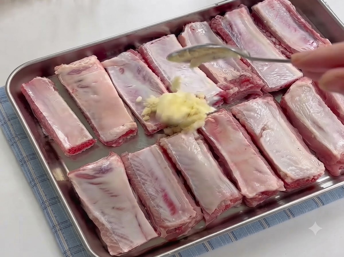 Una cuchara agregando un montón de ajo picado fresco sobre las costillas de cerdo crudas en una bandeja.