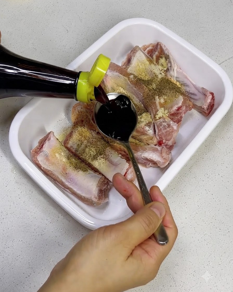 Vertiendo salsa de soya oscura desde una botella sobre costillas de cerdo crudas en una bandeja blanca.