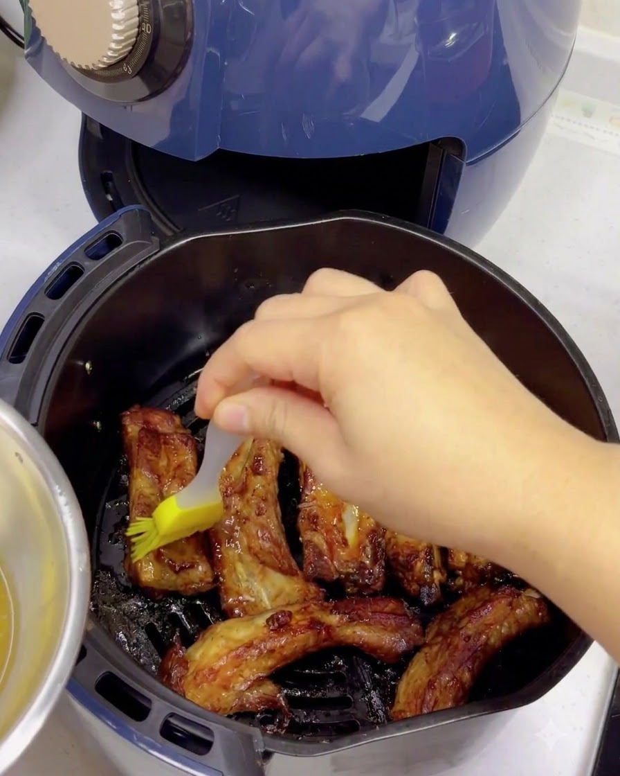 Un cepillo de silicona amarillo aplicando un glaseado de agua con miel sobre costillas de cerdo cocidas y doradas dentro de una canasta de freidora de aire.