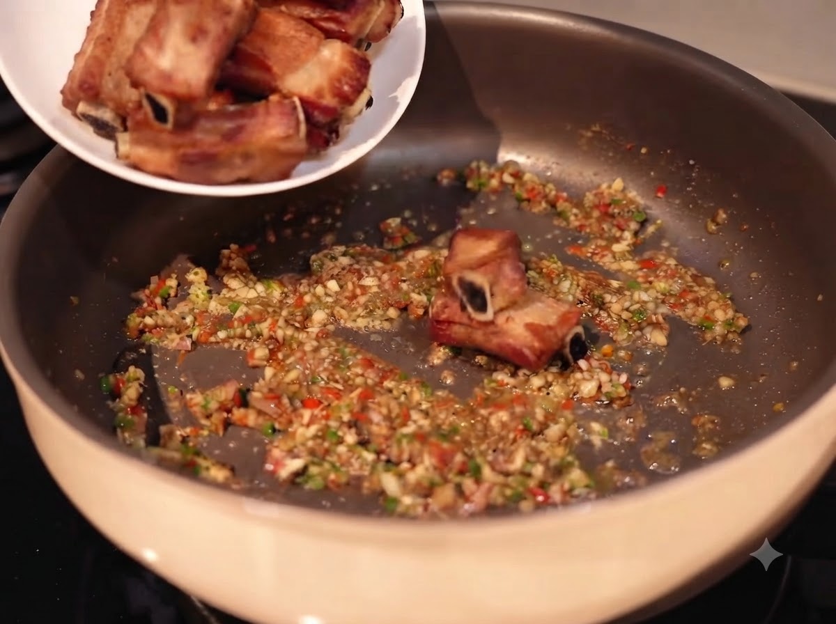 Costillas de cerdo siendo añadidas desde un plato blanco a una sartén llena de ajo picado salteado, chiles y especias.