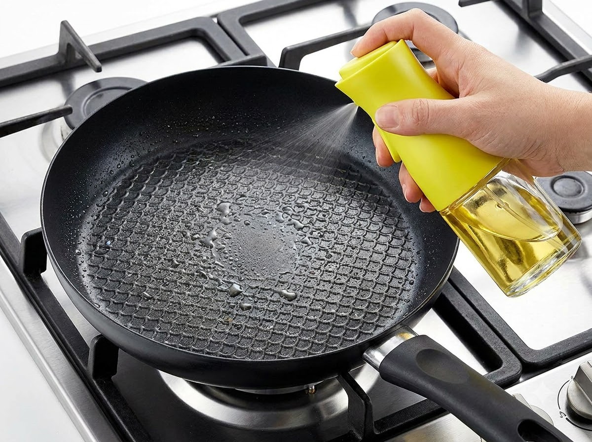Una mano rociando aceite de cocina en una sartén negra sobre un fogón encendido.