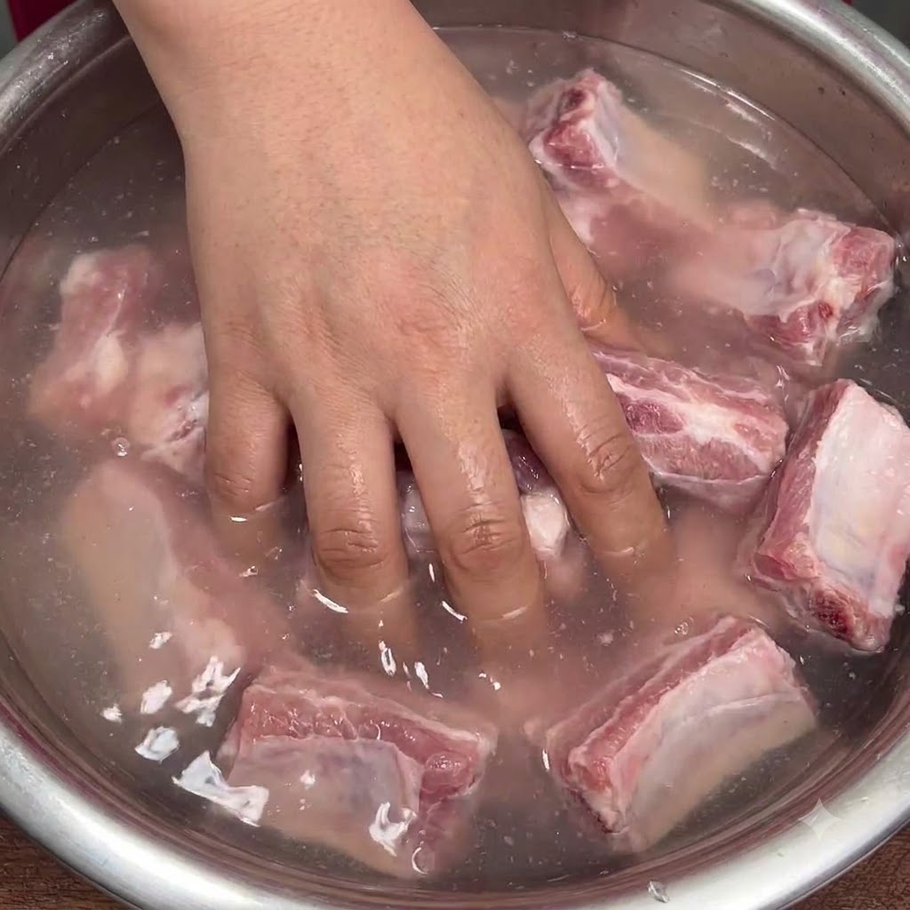 Una mano mezclando vigorosamente costillas de cerdo crudas en un tazón con agua turbia para distribuir el bicarbonato de sodio.