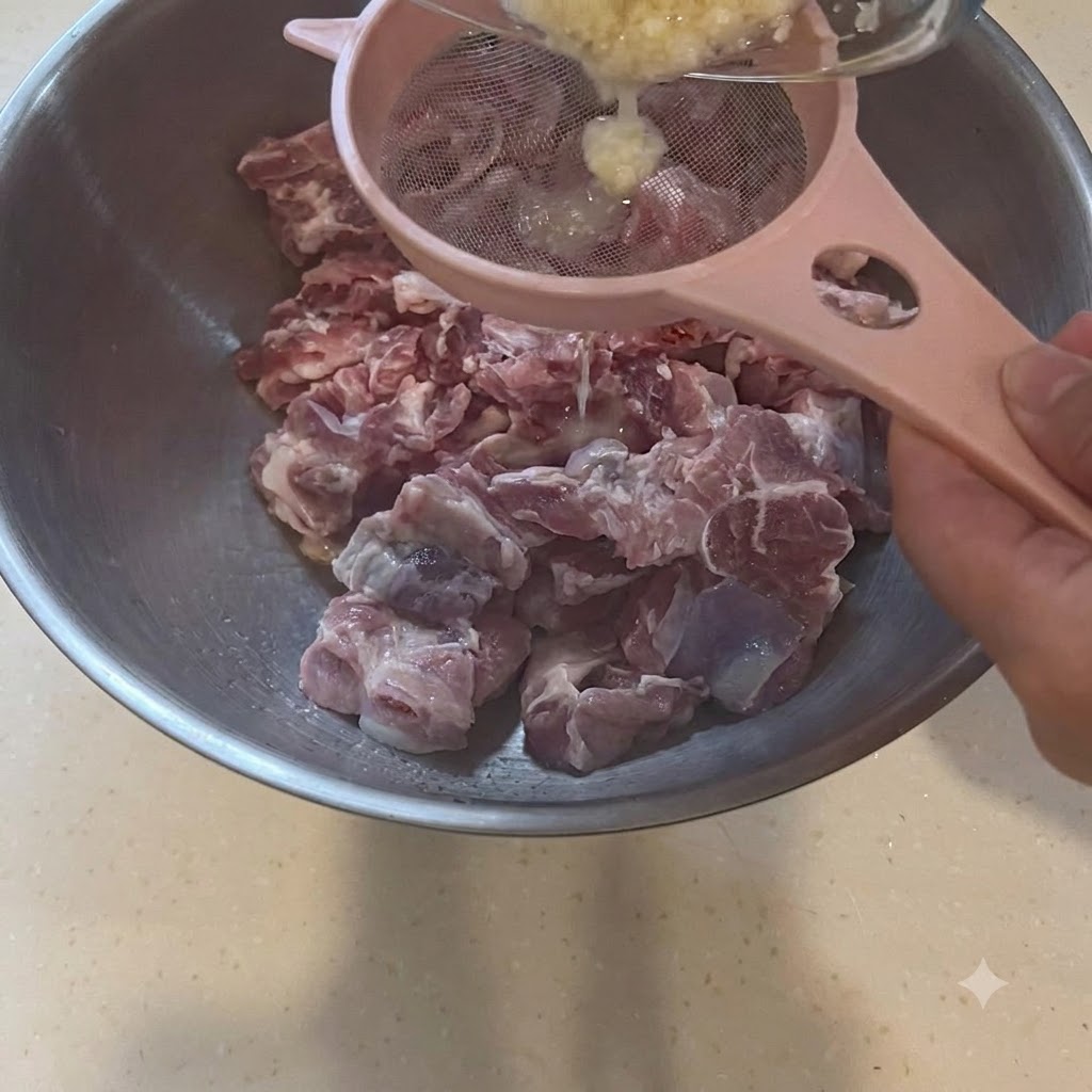 Pasta de ajo siendo colada a través de un colador rosa directamente sobre un bol grande con trozos de cerdo crudo.