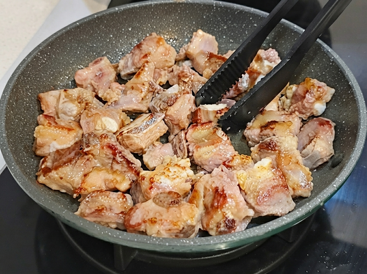 Costillas de cerdo siendo volteadas en una sartén antiadherente usando pinzas negras.